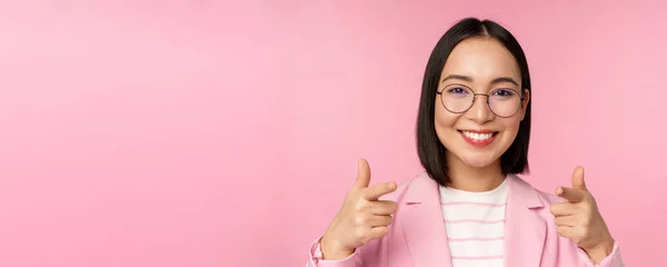 C'est toi, félicitations. Femme d'affaires asiatique souriante, PDG en costume et lunettes, pointant du doigt la caméra, recrutant, louant ou compliment, debout sur fond rose — Photo