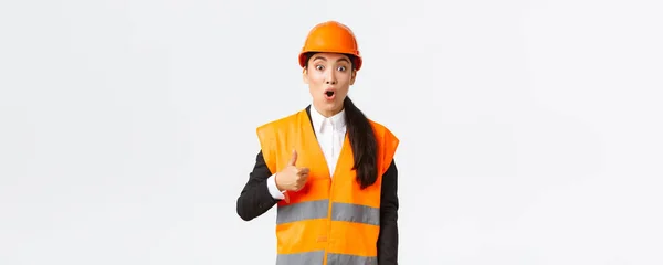 Ingénieur asiatique impressionnée et excitée en casque de sécurité et veste réfléchissante fascinée par un beau travail, montrant pouces levés haletant étonné, disons bien fait, excellent travail, debout fond blanc — Photo
