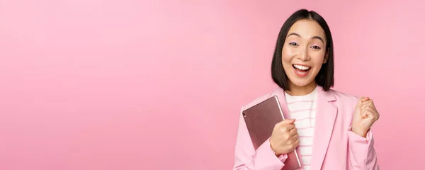 Entusiasta asiática empresária com tablet digital, gritando com alegria, triunfando, de pé sobre fundo rosa em terno — Fotografia de Stock