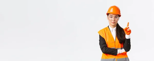 Спочатку безпека. Серйозна, усміхнена жінка азіатського інженера-будівельника, промислова жінка, що показує пальцем правила на підприємстві, носить захисні окуляри, шолом і рукавички, білий фон — стокове фото