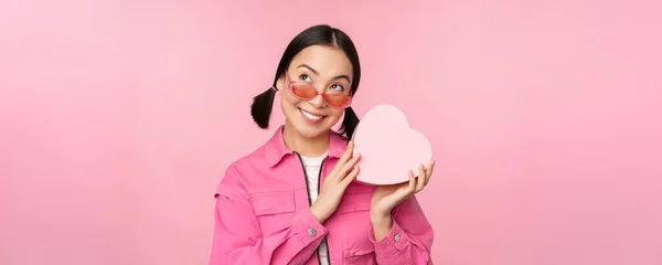 Imagem da namorada asiática elegante em óculos de sol, adivinhando o que está dentro da caixa de presente, presente em forma de coração, de pé sobre fundo rosa — Fotografia de Stock