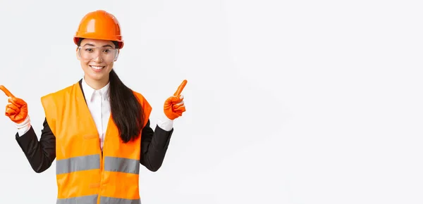 Sonriente profesional asiática mujer constructora, ingeniero en casco de seguridad, gafas de protección señalando los dedos hacia los lados, mostrando proyecto izquierdo y derecho, presentar proyectos de construcción, fondo blanco — Foto de Stock