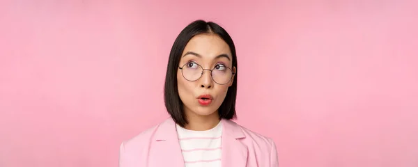 Lelkes eladónő, ázsiai üzletasszony szemüvegben, érdeklődik a bal felső sarok logója iránt, céges hirdetés, rózsaszín háttér fölött áll — Stock Fotó