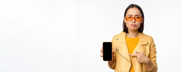 Retrato de mujer asiática triste en gafas de sol, apuntando con el dedo a la interfaz de la aplicación del teléfono móvil, mostrando la aplicación del teléfono inteligente, de pie sobre fondo blanco — Foto de Stock