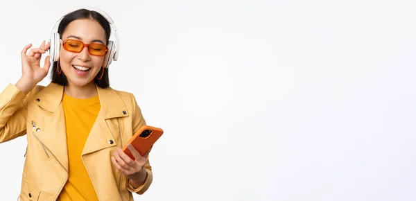 Felice donna asiatica elegante in occhiali da sole, ascoltare musica in cuffia, tenendo smartphone, canto e danza, in piedi su sfondo bianco — Foto Stock