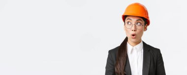 Etkilenmiş Asyalı kadın baş mimar, güvenlik kaskı ve iş elbisesi giymiş endüstriyel mühendis, şaşırmış ve şaşırmış görünüyor, fabrikada beyaz arka planda duruyor.