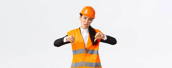 Desapontado e chateado asiático mulher engenheiro-chefe inspecionar mau trabalho, mostrando polegares para baixo decepcionado, usar capacete de segurança e reclamando fraco trabalho horrível do arquiteto, fundo branco — Fotografia de Stock
