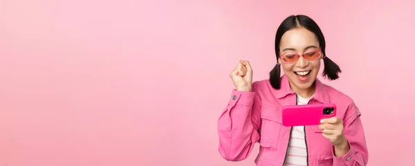 Menina coreana sorridente feliz ganhando no telefone celular, olhando para a tela horizontal do smartphone e alegrando-se, alcançar o objetivo, comemorando, de pé sobre o fundo rosa — Fotografia de Stock