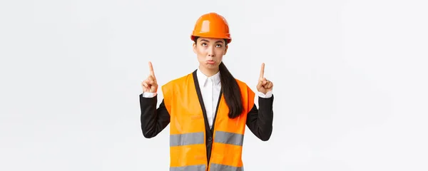 Épület, építőipar és ipari koncepció. Csalódott, komor ázsiai női mérnök, biztonsági sisakos építész és visszatükröződő ruházat duzzog elégedetlen, mert néz, és ujjal felfelé. — Stock Fotó