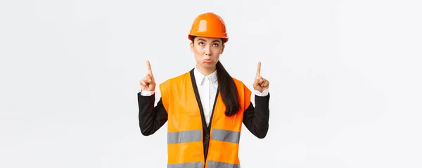 Construcción, construcción y concepto industrial. decepcionado sombrío asiático mujer ingeniero, arquitecto en seguridad casco y reflexivo ropa pucheo disgustado como mirando y señalando los dedos hacia arriba — Foto de Stock