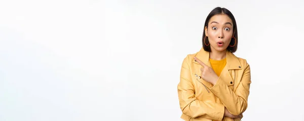 Retrato de hermosa mujer asiática expresar interés y emoción acerca de la publicidad, descuento tienda, señalando el dedo a la izquierda, mirando sorprendido, de pie sobre fondo blanco —  Fotos de Stock