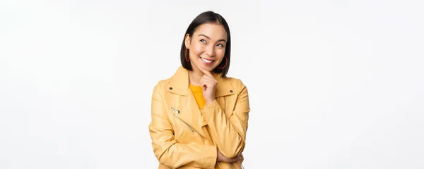 黄色のジャケットでスタイリッシュな韓国語の女性の肖像画、思慮深い笑顔、思考とロゴや広告を見上げ、白い背景の上に立って — ストック写真