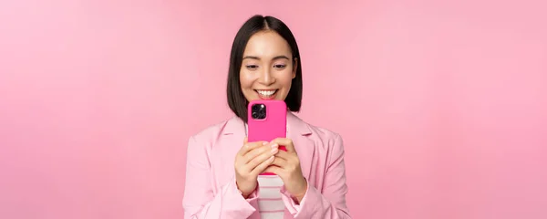 Imagem de mulher corporativa asiática sorridente em terno olhando, assistindo no aplicativo do smartphone, usando o aplicativo do telefone móvel, de pé sobre fundo rosa — Fotografia de Stock