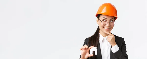 Creativo sorridente asiatico donna architetto pensando al nuovo design durante i lavori di costruzione, cercando premuroso con il sorriso come tenere piccola casa in miniatura, sfondo bianco — Foto Stock