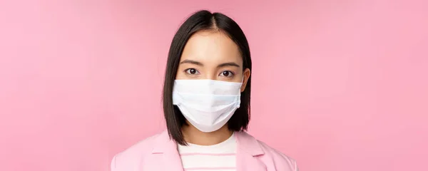 Nahaufnahme Porträt einer japanischen Geschäftsfrau mit medizinischer Gesichtsmaske, Anzug, Blick in die Kamera, vor rosa Hintergrund stehend — Stockfoto