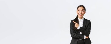 Heyecanlı ve eğlenceli Asyalı takım elbiseli genç iş kadını solu işaret ediyor ve beyaz arka planda duran reklam anonsuna heyecanlı bakıyor. Girişimci harika önerilerde bulunuyor.