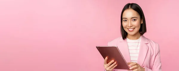 Porträt einer jungen asiatischen Corporate Frau, Bürodame mit digitalem Tablet, trägt Anzug, lächelt und sieht professionell aus, posiert vor rosa Hintergrund — Stockfoto