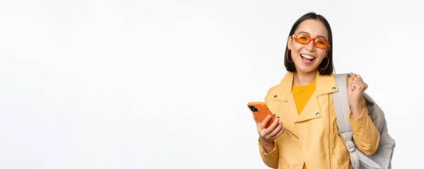 Ung asiatisk kvinna resenär, flicka turist i solglasögon, hålla ryggsäck och mobiltelefon, med hjälp av ansökan på smartphone, står över vit bakgrund — Stockfoto