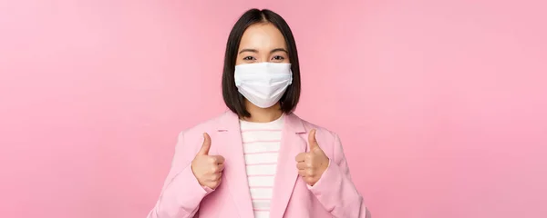 Empresária asiática de terno e máscara facial médica, mostrando os polegares para cima, recomendando o uso de equipamentos de proteção individual no escritório durante a pandemia covid-19, fundo rosa — Fotografia de Stock