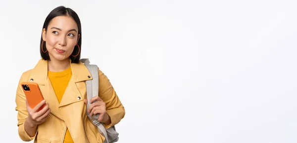 Азиатская девушка путешественник с рюкзаком, держа мобильный телефон, используя приложение смартфона, глядя вдумчивый, стоя на белом фоне — стоковое фото