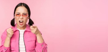 Image of angry, pissed off korean adult female model, shaking fists and shouting, screaming outraged, standing over pink background