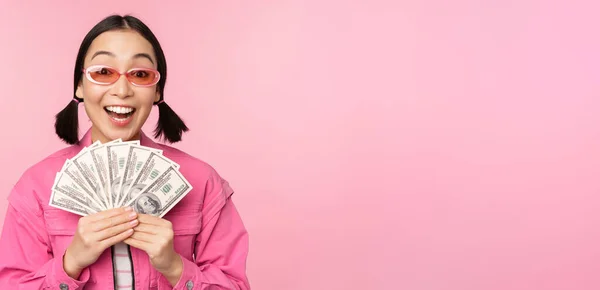 Conceito de microcrédito e empréstimos rápidos. Garota coreana elegante animado, mostrando dinheiro, dólares em dinheiro e olhando feliz, de pé em óculos de sol sobre fundo rosa — Fotografia de Stock