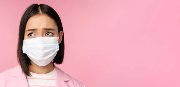 Close up retrato de triste, franzindo a testa asiática empresária em máscara facial médica, olhando para a esquerda no espaço cópia com decepcionado, emoção chateada, de pé sobre fundo rosa — Fotografia de Stock