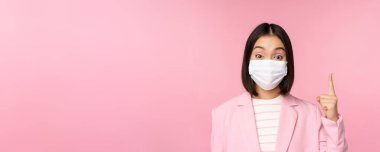 Tıbbi yüz maskesi ve takım elbiseli Asyalı iş kadını portresi, işaret ediyor, reklamını yapıyor, en üstte, pembe arka planda duruyor.