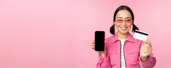 Bild av leende koreanska kvinna visar kreditkort och mobiltelefon skärm, smartphone applikationsgränssnitt, betala online, shopping kontaktlös, står över rosa bakgrund — Stockfoto