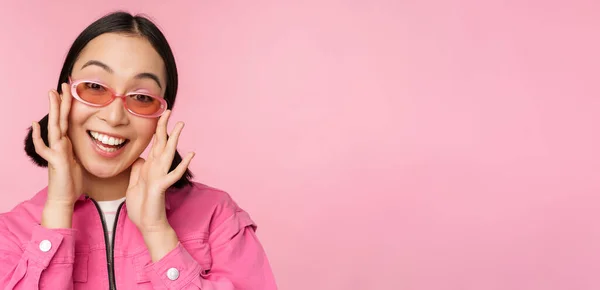 Χαριτωμένο σύγχρονο ιαπωνικό κορίτσι σε γυαλιά ηλίου, χαμογελώντας και αναζητούν ευτυχισμένη, ποζάροντας σε ροζ φόντο σε μοντέρνα ρούχα — Φωτογραφία Αρχείου