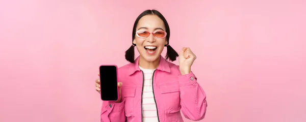 Возбужденная азиатка смеется и улыбается, показывает экран мобильного телефона, приложение для смартфона, стоя на розовом фоне — стоковое фото