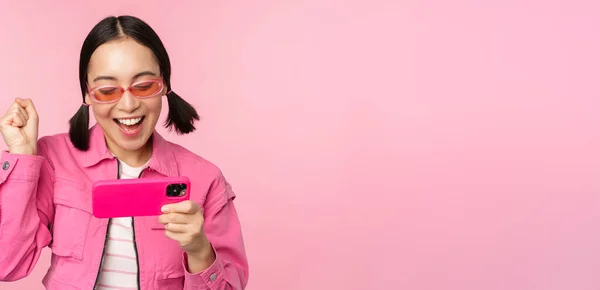 Щаслива дівчина, яка посміхається, виграє на мобільному телефоні, дивиться на горизонтальний екран смартфона і радіє, досягає мети, прославляє, стоїть над рожевим фоном. — стокове фото