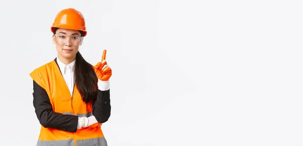 Veiligheid eerst. Serieuze, glimlachende vrouwelijke Aziatische bouwkundig ingenieur, industriële vrouw met de vinger naar het vertellen van regels over de onderneming, het dragen van een beschermende bril, helm en handschoenen, witte achtergrond — Stockfoto