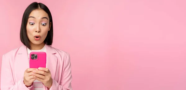 Porträtt av asiatisk affärskvinna med överraskad ansikte, med hjälp av smartphone app, bär kostym. Koreansk flicka med mobiltelefon och upphetsad ansikte uttryck, rosa bakgrund — Stockfoto