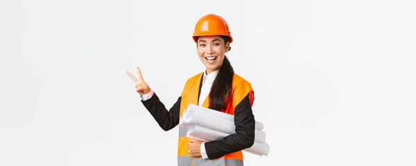 Glimlachend zelfverzekerd aziatische vrouwelijke ingenieur, bouwmanager in veiligheidshelm, dragen blauwdrukken, met twee vingers, zorgen voor het gebouw klaar in de tijd, staande witte achtergrond upbeat — Stockfoto