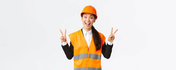 Joyeux sourire asiatique ingénieure, architecte en vêtements de sécurité et casque, montrant la paix signe kawaii et regardant optimiste, entreprise gagnant appel d'offres sur la construction et la construction, fond blanc — Photo