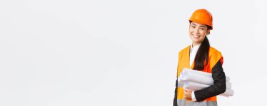 Gülümseyen başarılı Asyalı kadın mimar, güvenlik kasklı inşaat mühendisi, planlar taşıyor ve kameraya mutlu bir şekilde gülümsüyor, inşaat projesini tanıtıyor, beyaz arka planda duruyor.