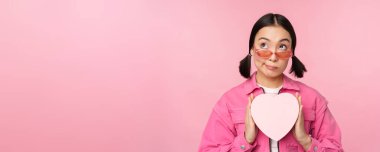 Güzel Asyalı kız mutlu gülümsüyor, kalp hediye kutusunu gösteriyor ve kameraya heyecanlı bakıyor, pembe romantik arka planda duruyor.