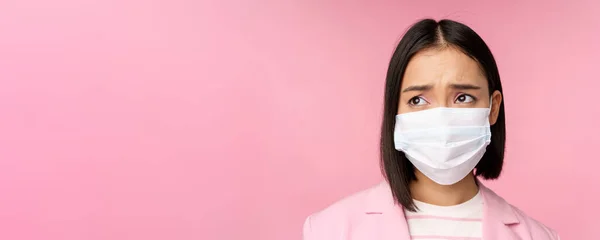 Portrait en gros plan d'une femme d'affaires asiatique triste et sournoise portant un masque médical, regardant à gauche l'espace de copie avec une émotion déçue et bouleversée, debout sur un fond rose — Photo