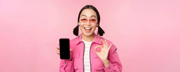 Entuzjastyczna młoda azjatycka kobieta pokazuje w porządku, ok znak, uśmiechnięty zadowolony, ekran telefonu komórkowego, aplikacja smartfona, stojąc na różowym tle — Zdjęcie stockowe
