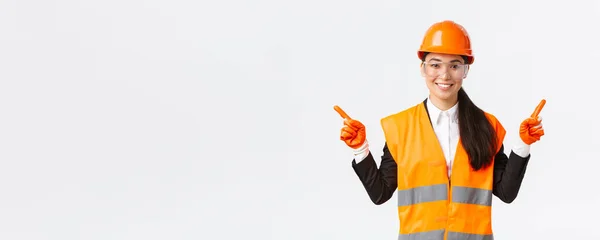 Усміхнена професійна азіатська жінка-будівельник, інженер в шоломі безпеки, захисні окуляри спрямовані пальцями вбік, показують лівий і правий проект, вводять будівельні проекти, білий фон — стокове фото