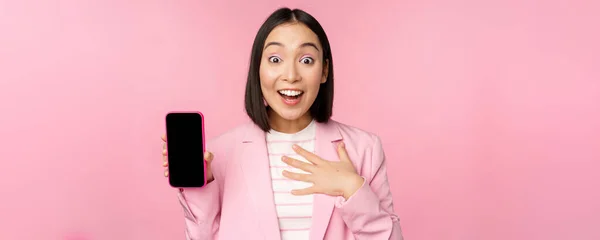 Удивлена, восторженный азиатский бизнесмен показывает экран мобильного телефона, смартфон интерфейс приложения, стоя на розовом фоне — стоковое фото