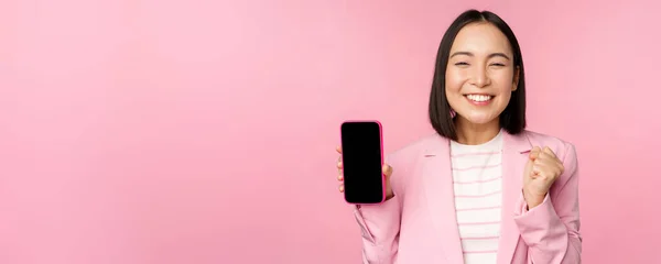Vinnande koreanska affärskvinna visar smartphone skärm, ler nöjd, demonstrera mobiltelefon ansökan, webbutik eller shopping app, står över rosa bakgrund — Stockfoto