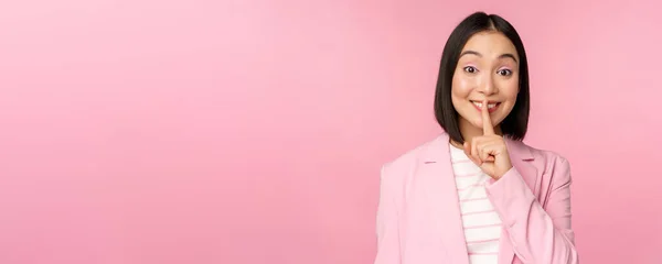 Tiše, tabu. Portrét asijské podnikatelky ukazující shush gesto, shhh znamení, stiskněte prst na rty, stojící nad růžovým pozadím v obleku — Stock fotografie