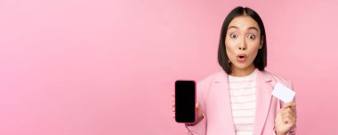 Ateşli Asyalı iş kadını cep telefonu ekranı ve kredi kartı gösteriyor, kameraya hayran bakıyor, pembe arka planda duruyor.