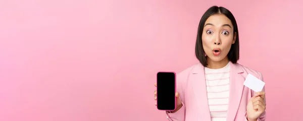 Entusiástica asiática empresária mostrando tela do telefone móvel e cartão de crédito, olhando espantado para a câmera, de pé sobre fundo rosa — Fotografia de Stock