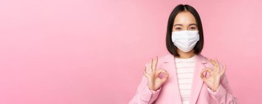 Takım elbiseli ve sağlık maskeli Asyalı bir iş kadını, tamam işareti gösteriyor, covid-19 salgını sırasında koruyucu ekipman kullanmayı öneriyor, pembe arka plan.