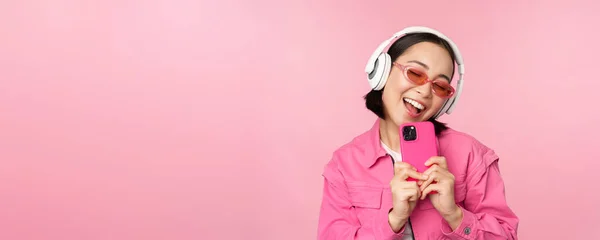 Chica asiática con estilo en los auriculares, escuchar música y tomar fotos en el teléfono móvil, utilizando el teléfono inteligente, de pie sobre el fondo rosa — Foto de Stock