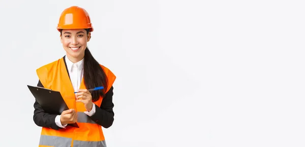 Confiado sonriente mujer asiática construcción ingeniero, industrial mujer en seguridad casco visita edificio área para inspección, anotando notas en portapapeles y buscando satisfecho, fondo blanco — Foto de Stock