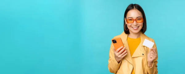 Online shopping. Stylish young asian woman in sunglasses, showing credit card and using smartphone, paying in internet, making purchase, standing over blue background — Stock Photo, Image